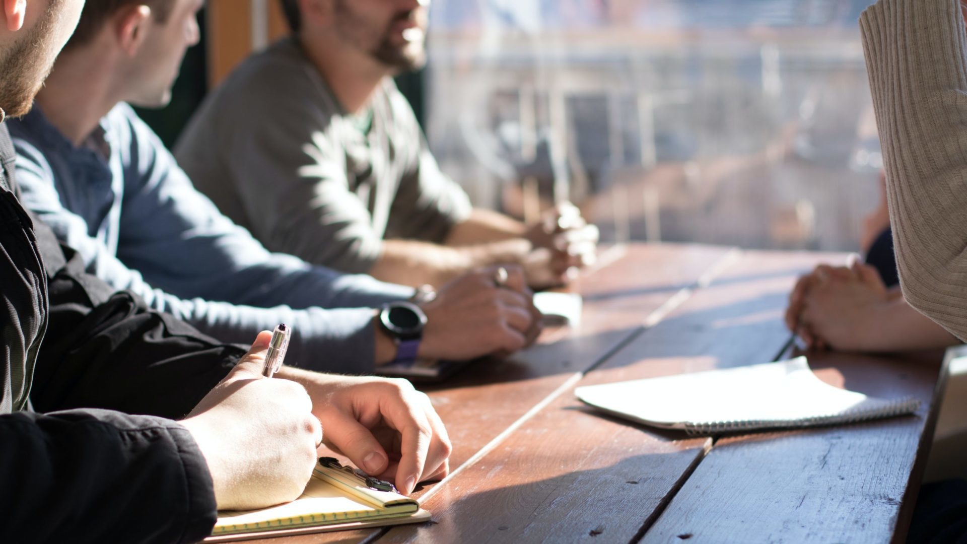 Meeting di 5 persone attorno a un tavolo di legno, con focus su una mano che prende appunti su un quaderno