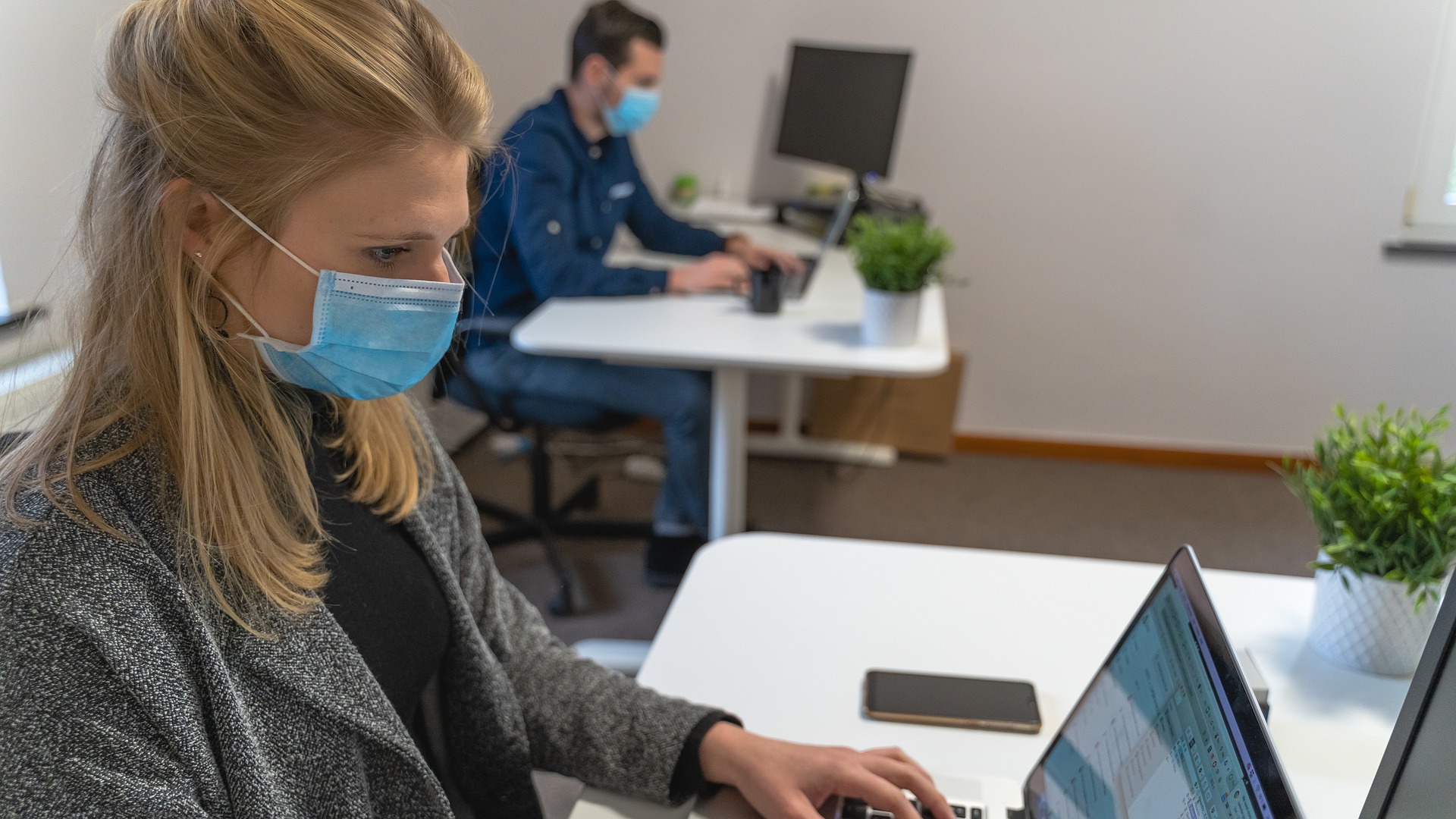 ragazza a lavoro a una scrivania del suo ufficio che indossa la mascherina e scrive al computer