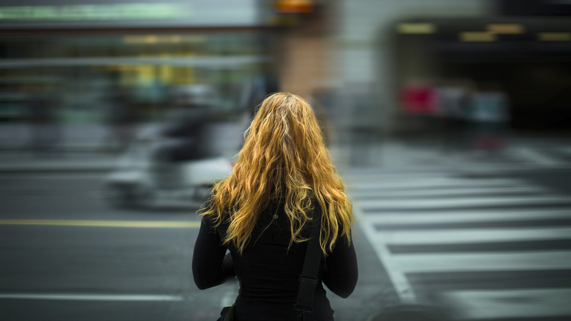 ragazza bionda mezzo busto e di spalle che sta attraversando una strada in prossimità delle strisce pedonali