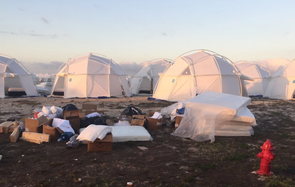 Su una spiaggia piena di spazzatura, sullo sfondo si vedono delle tende bianche al tramonto